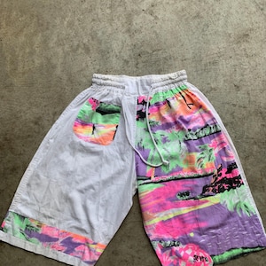 Puede incluir: Pantalones cortos blancos con un estampado tropical colorido en un lado y un bolsillo. El estampado presenta palmeras, una puesta de sol y el texto "SEV YD" en rosa. Los pantalones cortos tienen cintura elástica y cordón.