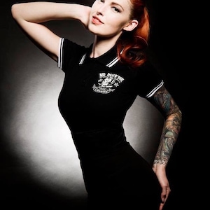 May include: A black polo shirt with white trim and the text "MR. DUCKTAIL LONDON" is worn by a person with red hair. The shirt has a logo on the chest. The person has a tattoo sleeve on their arm.