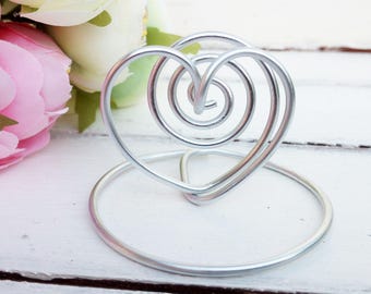May include: Silver wire heart shaped photo holder. The heart is made of two intertwined spirals. The holder is on a white wooden surface.