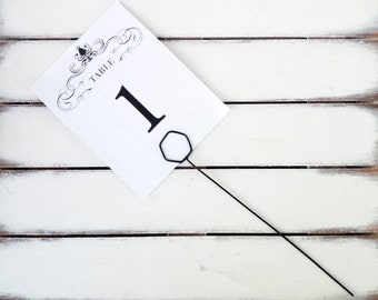 May include: White wooden table number holder with a black metal rod and a white card with black text that reads "Table 1".