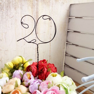 Può includere: Un cake topper nero a filo metallico con il numero 20 e un design a ricciolo, posto sopra una colorata composizione floreale. I fiori includono fiori rossi, gialli, rosa e bianchi, creando un'esposizione vibrante. Lo sfondo presenta un muro usurato e un elemento decorativo bianco.