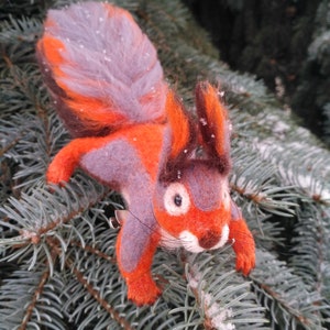 May include: A hand-felted squirrel toy with orange and gray fur, sitting on a snowy evergreen branch.