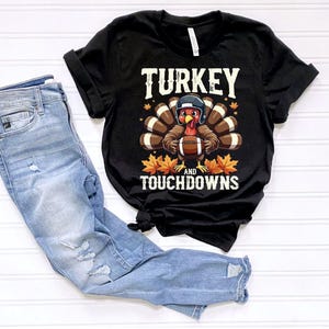May include: Black t-shirt with the text "TURKEY and TOUCHDOWNS" and a cartoon turkey wearing a football helmet and holding a football. Light blue denim jeans are next to the shirt. The shirt has rolled-up sleeves.
