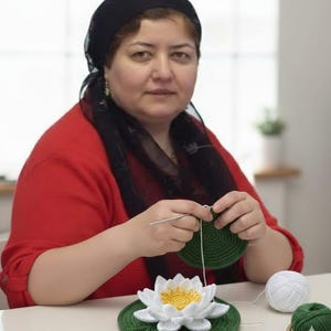 May include: A handmade crochet water lily with white petals and a yellow center, resting on a green lily pad. A woman is crocheting a green circle, with white and green yarn nearby. The woman is wearing a red shirt and a black head covering.