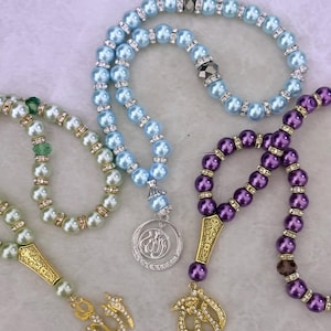 Op de afbeelding: Drie gebedskettingen met kralen en gouden bedels. De kettingen zijn lichtblauw, groen en paars. De bedels zijn gevormd als het Arabische woord voor God, "Allah".