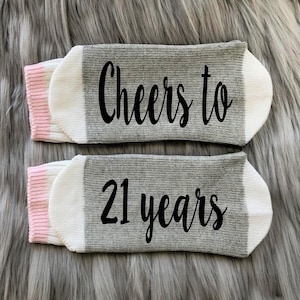 May include: A pair of gray and white socks with pink trim. The socks have the text "Cheers to 21 years" printed on them.