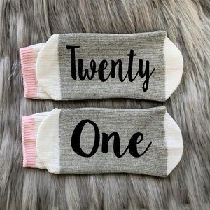 May include: A pair of gray and white socks with pink trim. The socks have the words "Twenty One" written on them in black.