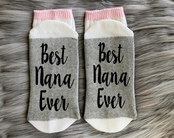 May include: A pair of gray socks with the words "Best Nana Ever" in black script. The socks have white foot and heel sections, and pink and white striped cuffs. The socks are displayed on a gray, furry surface.