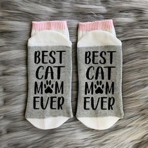 May include: A pair of grey and white socks with a pink stripe at the top. The socks have the text "Best Cat Mom Ever" printed on them in black.