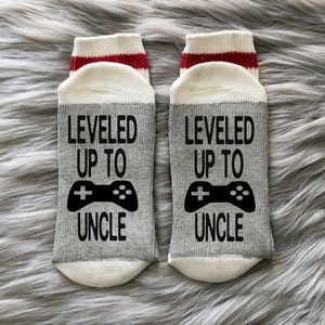 May include: A pair of gray and white socks with red stripes at the top. The socks have a black and white video game controller graphic and the text "LEVELED UP TO UNCLE".