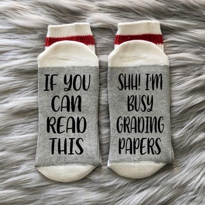 May include: A pair of gray and white socks with red stripes. One sock says "If you can read this" and the other sock says "Shh! I'm busy grading papers".