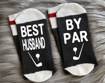 May include: A pair of dark gray socks with white text that reads "BEST HUSBAND" and "BY PAR". Each sock features a golf club and ball graphic. The socks have white toes and cuffs with red stripes.