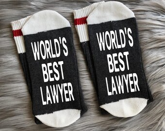 May include: A pair of black socks with white and red stripes. The socks have the text "World's Best Lawyer" printed on them in white.