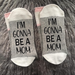 Op de afbeelding: Een paar grijze en witte sokken met roze strepen. E&eacute;n sok zegt "Ik ga mama worden" en de andere sok zegt "Ik ga mama worden".