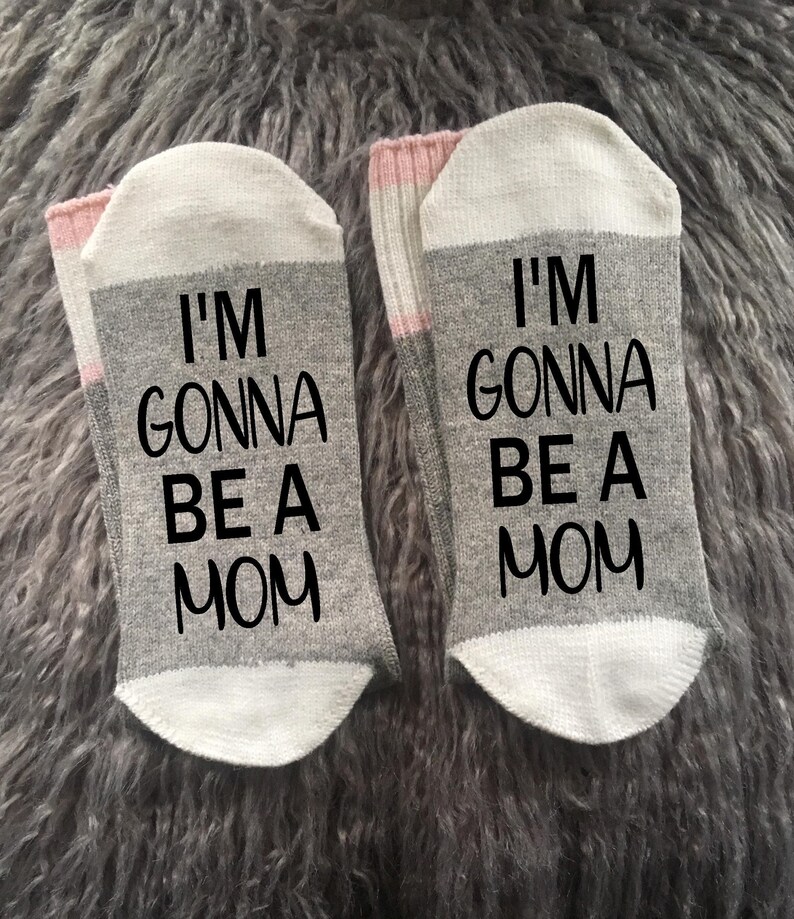 Op de afbeelding: Een paar grijze en witte sokken met roze strepen. E&eacute;n sok zegt "Ik ga mama worden" en de andere sok zegt "Ik ga mama worden".