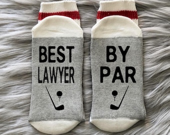 May include: A pair of gray and white socks with red trim. One sock says "BEST LAWYER" with a golf club and ball graphic. The other sock says "BY PAR" with a golf club and ball graphic.