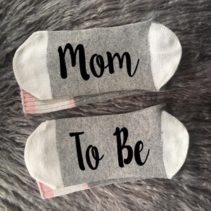 May include: A pair of gray socks with white trim. One sock says "Mom" and the other says "To Be".