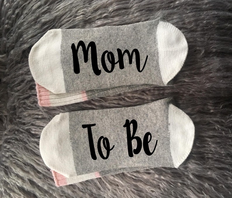 May include: A pair of gray socks with white trim. One sock says "Mom" and the other says "To Be".