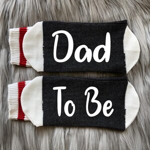 May include: A pair of black socks with white trim and red stripes. One sock says "Dad" and the other says "To Be".