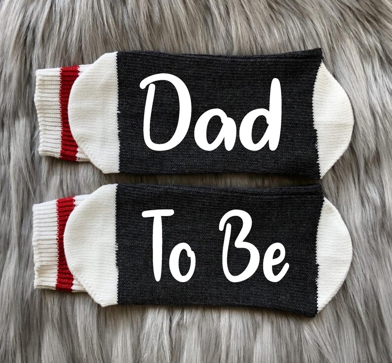 May include: A pair of black socks with white trim and red stripes. One sock says "Dad" and the other says "To Be".