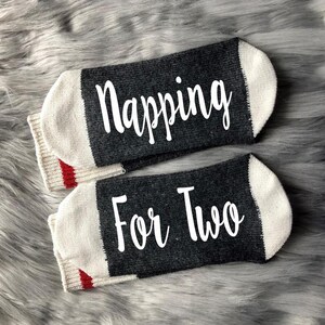 Op de afbeelding: Een paar grijze en witte sokken met rode bies. E&eacute;n sok zegt "Napping" en de andere zegt "For Two".