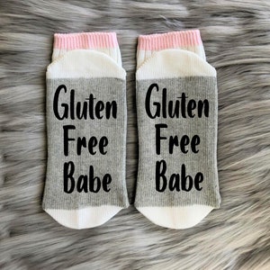 K&ouml;nnte beinhalten: Ein Paar graue und wei&szlig;e Socken mit einem rosa Streifen oben. Die Socken haben den Text "Gluten Free Babe" in Schwarz aufgedruckt.