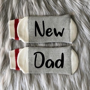May include: A pair of gray socks with white cuffs and red stripes. One sock says "New" and the other says "Dad".