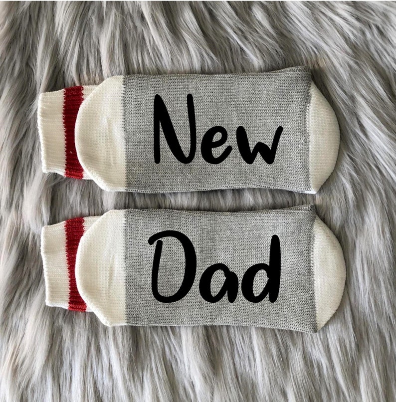 May include: A pair of gray socks with white cuffs and red stripes. One sock says "New" and the other says "Dad".