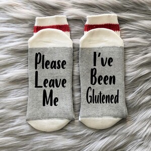 May include: A pair of gray and white socks with red stripes. One sock says "Please Leave Me" and the other says "I've Been Glutened".