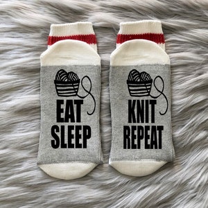 Puede incluir: Un par de calcetines grises con punteras y talones blancos, y pu&ntilde;os a rayas rojas y blancas. Los calcetines presentan un gr&aacute;fico negro de una cesta de hilo y las palabras "EAT SLEEP KNIT REPEAT" en letras negras.