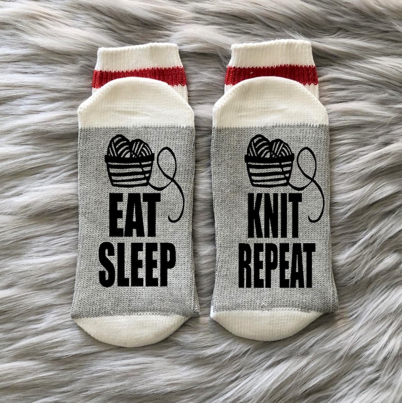 Puede incluir: Un par de calcetines grises con punteras y talones blancos, y pu&ntilde;os a rayas rojas y blancas. Los calcetines presentan un gr&aacute;fico negro de una cesta de hilo y las palabras "EAT SLEEP KNIT REPEAT" en letras negras.