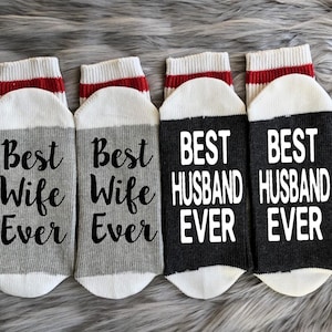 Puede incluir: Cuatro pares de calcetines con rayas rojas y blancas en la parte superior. Dos pares son grises con el texto "Best Wife Ever" en negro. Dos pares son negros con el texto "Best Husband Ever" en blanco.