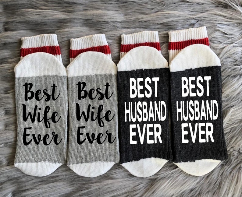 Puede incluir: Cuatro pares de calcetines con rayas rojas y blancas en la parte superior. Dos pares son grises con el texto "Best Wife Ever" en negro. Dos pares son negros con el texto "Best Husband Ever" en blanco.