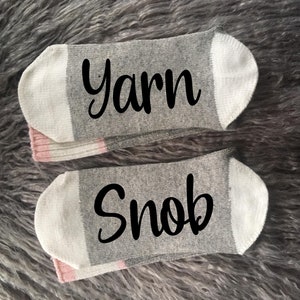 Puede incluir: Dos calcetines tobilleros grises con punteras y talones blancos y detalles rosas. Un calcet&iacute;n tiene la palabra "Yarn" en escritura negra, y el otro dice "Snob". Los calcetines se muestran sobre una superficie gris texturizada.
