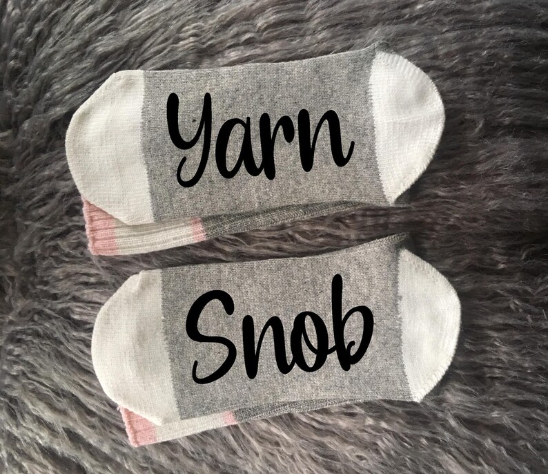 Puede incluir: Dos calcetines tobilleros grises con punteras y talones blancos y detalles rosas. Un calcet&iacute;n tiene la palabra "Yarn" en escritura negra, y el otro dice "Snob". Los calcetines se muestran sobre una superficie gris texturizada.