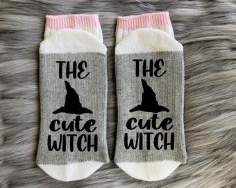 May include: A pair of gray and white socks with a pink trim. The socks have a black witch hat and the text "The cute witch" printed on them.