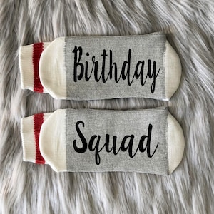 Puede incluir: Un par de calcetines grises con pu&ntilde;os blancos y rayas rojas. Un calcet&iacute;n dice "Birthday" y el otro dice "Squad".
