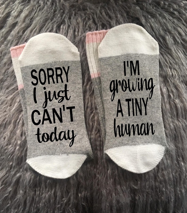 Op de afbeelding: Een paar grijze en witte sokken met roze accenten. E&eacute;n sok heeft de tekst "Sorry, ik kan het vandaag gewoon niet" en de andere "Ik kweek een klein mensje".