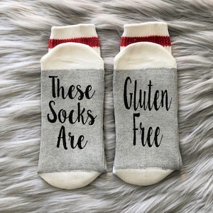 K&ouml;nnte beinhalten: Ein Paar graue und wei&szlig;e Socken mit rotem Besatz. Eine Socke sagt "These Socks Are" und die andere Socke sagt "Gluten Free".