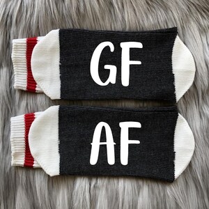 May include: A pair of black socks with white lettering. One sock says "GF" and the other says "AF". The socks have a white cuff with a red stripe.