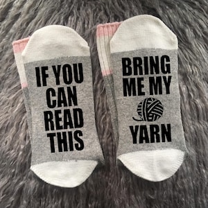 Puede incluir: Un par de calcetines grises con punteras y pu&ntilde;os blancos, y una banda rosa. Un calcet&iacute;n dice "IF YOU CAN READ THIS" en texto negro, y el otro dice "BRING ME MY YARN" con un gr&aacute;fico de ovillo.