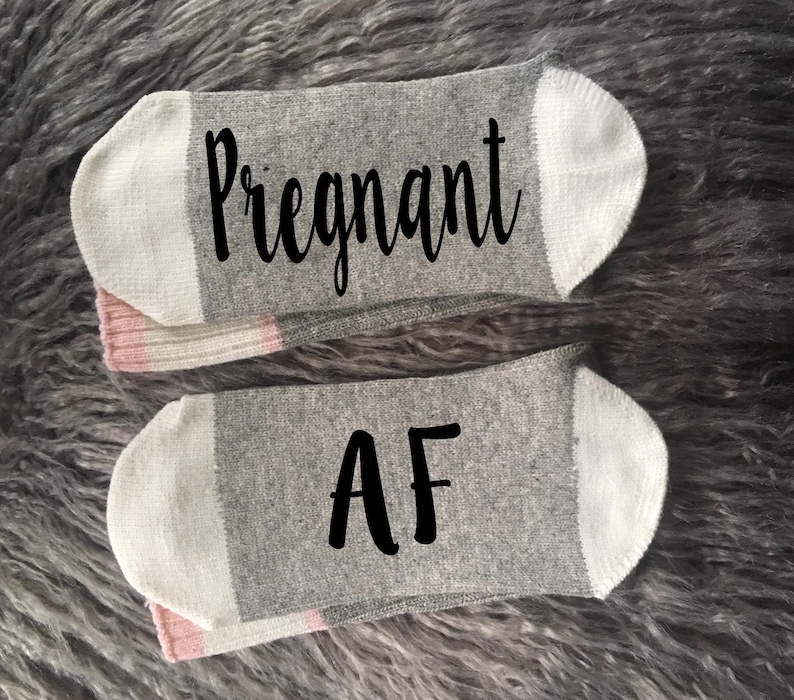Op de afbeelding: Een paar grijze en witte sokken met roze bies. De sokken hebben de tekst "Pregnant AF" erop gedrukt.