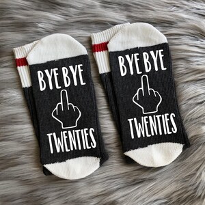 May include: A pair of black socks with white lettering that says "Bye Bye Twenties" with a middle finger emoji. The socks have a white cuff with a red stripe.