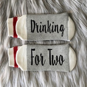 Op de afbeelding: Een paar grijze en witte sokken met rode strepen. E&eacute;n sok zegt "Drinking" en de andere zegt "For Two".