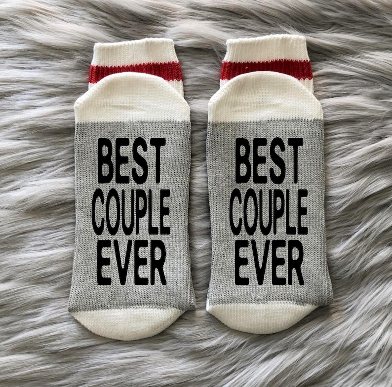 Puede incluir: Un par de calcetines grises y blancos con rayas rojas. Los calcetines tienen el texto "Best Couple Ever" impreso en negro.