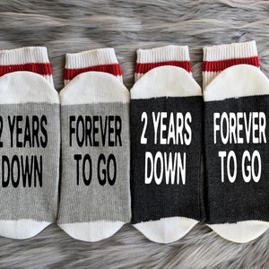Puede incluir: Cuatro pares de calcetines grises y negros con rayas rojas. Los calcetines tienen el texto "2 Years Down" y "Forever To Go" impreso en ellos.