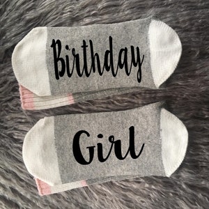 May include: A pair of gray socks with pink stripes. One sock says "Birthday" and the other says "Girl".