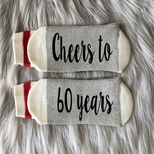 Puede incluir: Un par de calcetines grises y blancos con rayas rojas. Los calcetines tienen el texto "Cheers to 60 years" impreso en ellos.