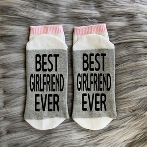 Op de afbeelding: Een paar grijze en witte sokken met een roze streep aan de bovenkant. De sokken hebben de tekst "BEST GIRLFRIEND EVER" in zwart op het grijze gedeelte gedrukt.