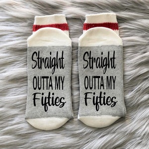 Puede incluir: Un par de calcetines blancos hasta el tobillo con una franja roja en la parte superior. Los calcetines tienen una franja gris en el medio con el texto "Straight outta my fifties" impreso en negro.